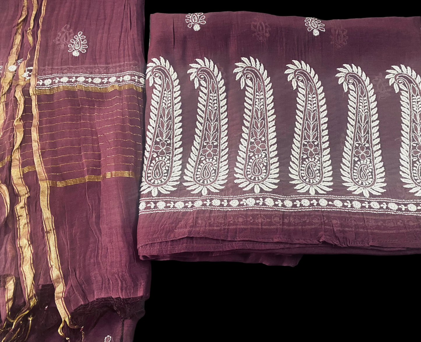 A folded piece of deep maroon fabric with white chikankari embroidered paisley motifs and gold striped border details, shown alongside another matching textile on a dark green surface.